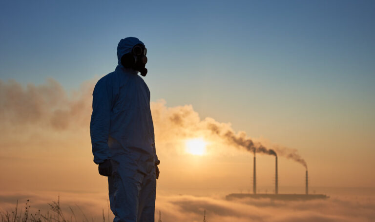 Scientist wearing a white coverall and gas mask standing bfore smoking pipes of thermal power plant