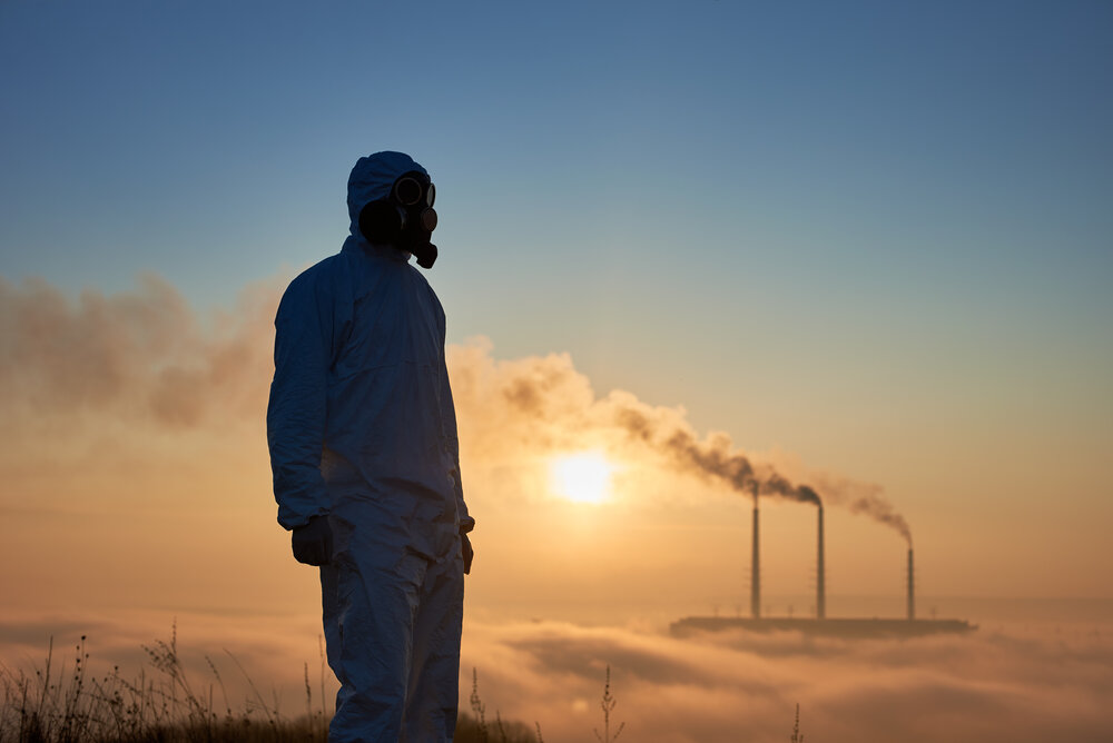 Scientist wearing a white coverall and gas mask standing bfore smoking pipes of thermal power plant
